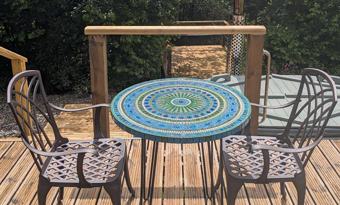 Handmade mosaic table in blue and green with metal dining armchairs on a wood deck with the hot tub, trees and lake in the background.